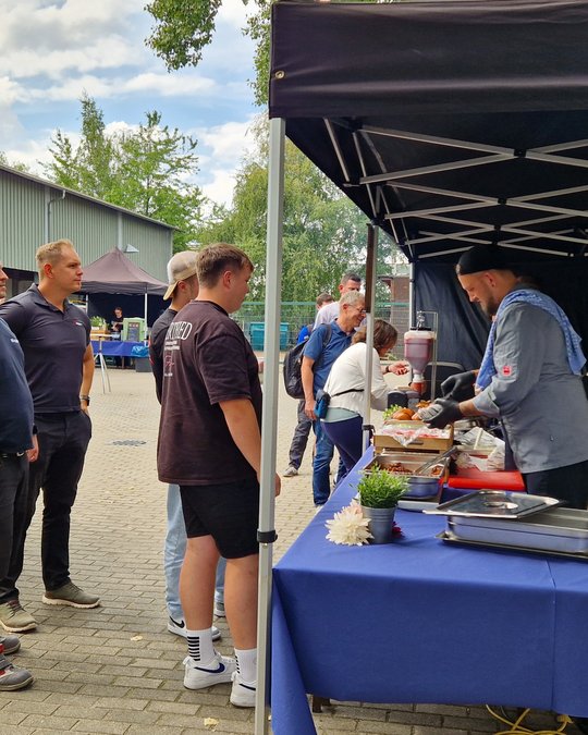 Eine Gruppe von Menschen steht in der Schlange an einem Verkaufsstand, wo ein Koch Speisen zubereitet. Der Stand ist mit blauem Tuch bedeckt, und es sind verschiedene Gerichte auf dem Tisch zu sehen. Im Hintergrund sind weitere Besucher und ein sonniger Himmel.