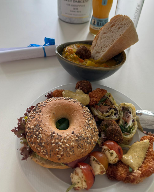 A plate featuring a sesame bagel with lettuce, several rolled wraps, falafel, and cherry tomatoes drizzled with sauce. A bowl of soup with vegetables and a piece of bread accompany the dish. Various condiments and utensils are present on the table.