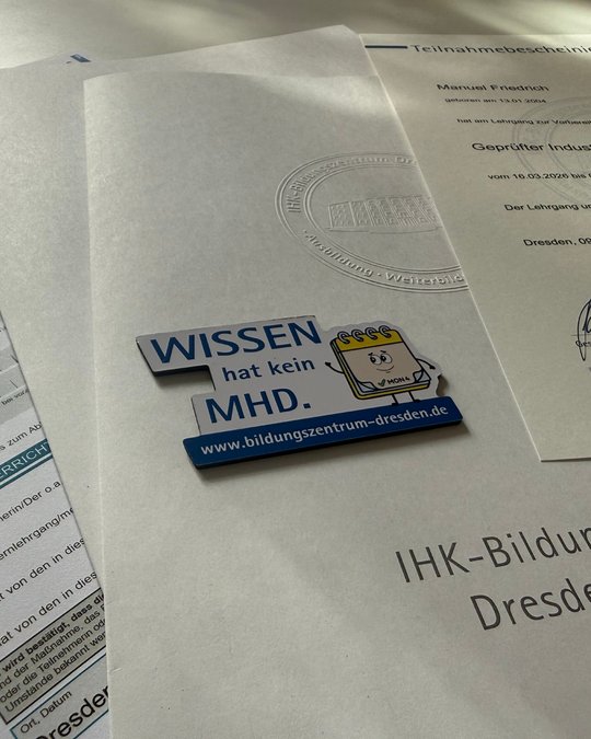 A close-up image of several papers including a participation certificate and informational documents, with a blue and white promotional magnet that reads "WISSEN hat kein MHD" with a cartoon character, placed prominently on top.