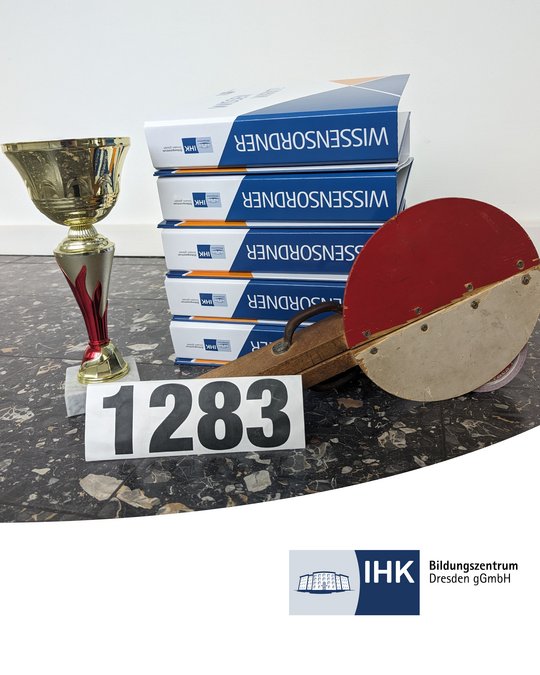 Eine goldene Trophäe steht neben einer Holzschnittschnalle und einer Nummerntafel mit der Aufschrift „1283“. Im Hintergrund sind Stapel von Broschüren mit dem Titel „Wissensordner“ sichtbar, die zur IHK Bildungszentrum Dresden gehören.