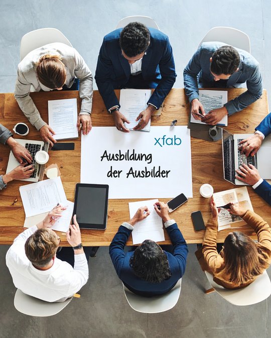Eine Gruppe von sechs Personen sitzt an einem Holztisch, umgeben von Laptops und Unterlagen. Im Mittelpunkt liegt ein Blatt mit der Aufschrift "Ausbildung der Ausbilder" und dem Logo von xfab. Die Teilnehmer sind in angeregter Diskussion vertieft.