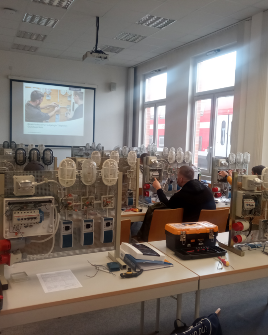 The image shows a classroom setting with multiple tables set up for practical training. Devices and equipment are arranged on the tables, while participants engage with the materials. A projector displays educational content on a screen, illuminating the collaborative learning environment.