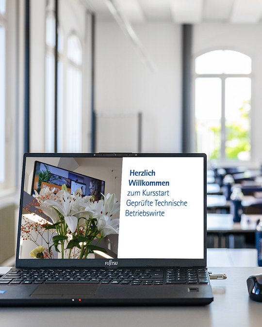 A laptop sits on a table displaying a welcome message in German, announcing the start of a course for certified technical business administrators. In the background, a bright classroom is visible with rows of desks and large windows. A vase with flowers is also shown on the screen.