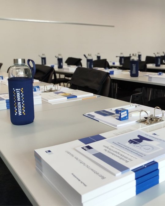 Auf einem Tisch für eine Veranstaltung sind mehrere Stapel von Informationsmaterialien und ein Getränkebehälter zu sehen. Der Getränkebehälter ist in Blau gehalten und trägt die Aufschrift „Wissen wirkt!“. Der Raum wirkt vorbereitet für eine Schulung oder ein Seminar.
