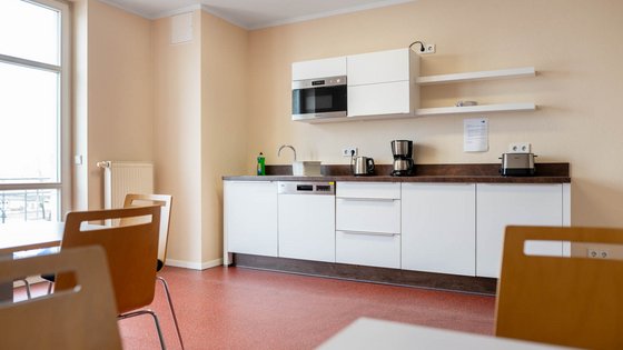 A modern kitchen with white cabinets and a brown countertop. It features a microwave, coffee maker, and kettle on the countertop. A green bottle is visible, and a few chairs are positioned in front of a table. Sunlight enters through a window.