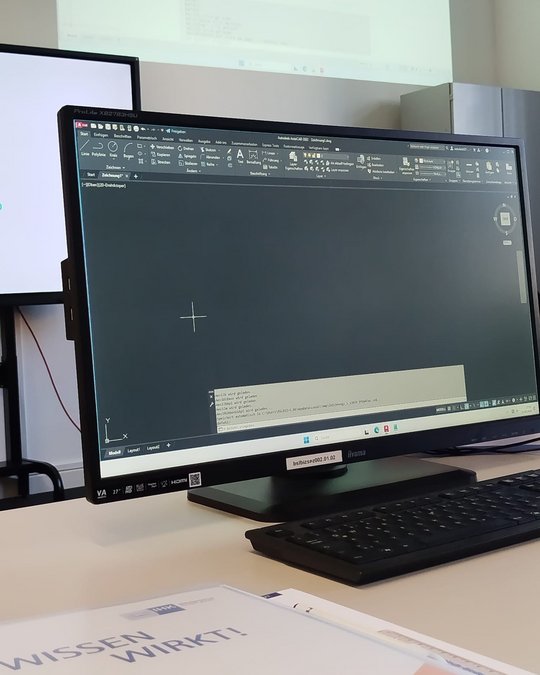 A computer monitor displaying a dark background with computer-aided design (CAD) software open. A command line and coordinate crosshair are visible. In the foreground, a black keyboard rests on the table, and a document partially shows the title “WISSEN WIRKT!” in the lower part of the image.