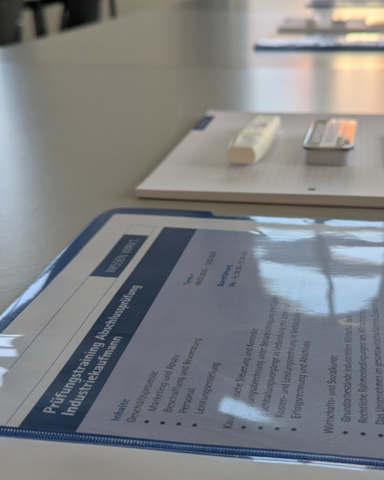 A close-up view of a neatly arranged table featuring a document in a plastic cover, a notepad, and a pen. The document is titled "Prüfungsprotokoll" and pertains to an industrial examination. The surface of the table reflects natural light.