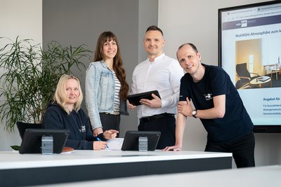 Four professionals are gathered around a modern workspace. They are engaged in a discussion, with two members seated at a table and two standing beside them. A large screen displays an interior design project in the background, and a plant adds a touch of greenery to the setting.