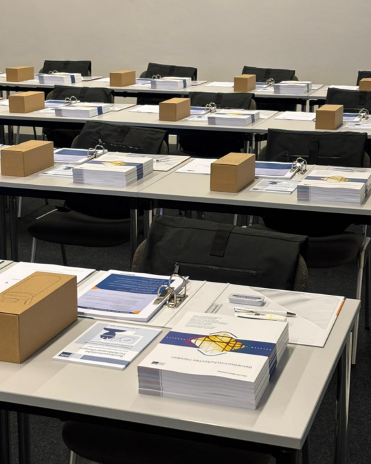 A classroom setting featuring several tables arranged with materials for a meeting or seminar. Each table has stacks of paper, binders, pens, and small brown boxes. The environment is organized and prepared for participants, with empty chairs indicating that attendees are not yet present.