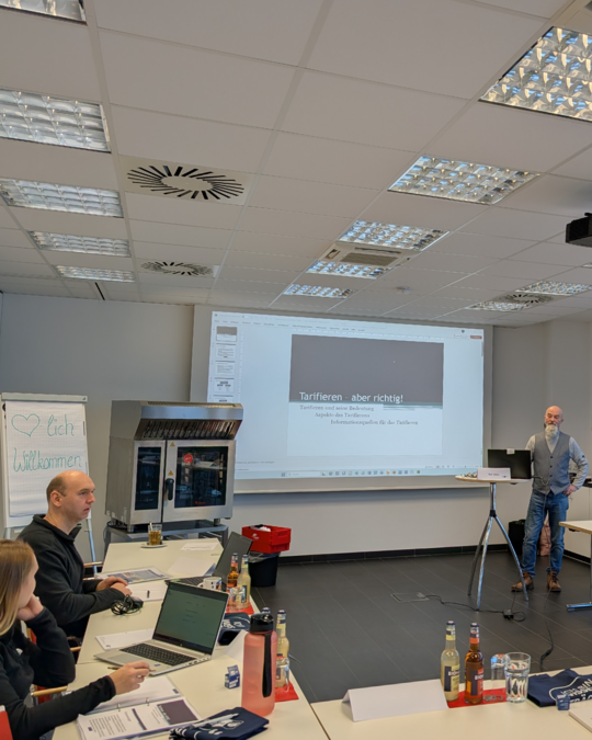 A seminar room with participants seated at a table, focusing on a presentation displayed on a screen. The slide titled "Tarifieren - aber richtig!" highlights a discussion about proper pricing strategies. A presenter stands near the screen, while laptops and beverages are visible on the table.