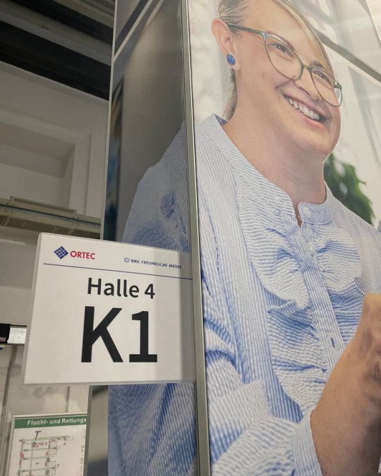 A sign labeled "Halle 4 K1" is displayed on a wall next to a smiling person with glasses, wearing a blue-striped shirt. The image conveys a welcoming atmosphere, likely in an exhibition or event space.