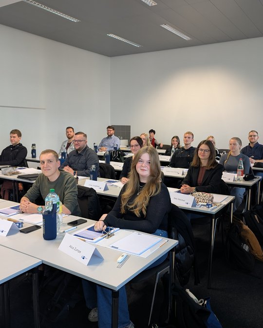 A group of students is seated in a classroom, attentively looking toward the front. Desks are organized with notebooks and water bottles. The atmosphere is focused and engaged, with several students smiling. The setting suggests a learning environment.
