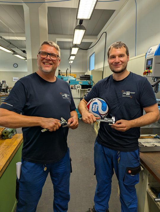 Two men smile while standing in a workshop. They hold adjustable wrenches in one hand and a soccer ball in the other. The background features machines and workstations, indicating a technical environment. Both men are wearing navy blue shirts and blue pants.