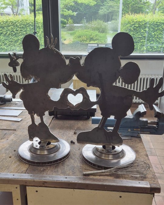 Zwei metallene Silhouetten von Mickey und Minnie Mouse küssen sich. Zwischen ihnen befindet sich ein Herz. Die Figuren stehen auf runden, stabilen Sockeln und sind auf einer Holzfläche platziert, im Hintergrund sind grüne Bäume sichtbar.