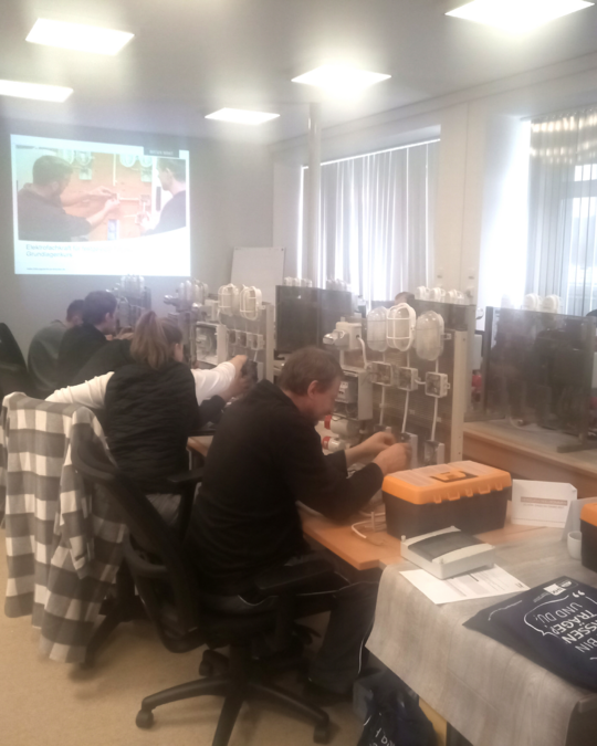 A training session is taking place in a classroom setting. Participants are seated at desks, working with equipment displayed on shelves. A projector screen shows instructional material in the background. The environment is bright and organized, with various tools and materials visible.